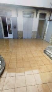 Patio extérieur carrelé marron clair, partiellement mouillé, devant une façade blanche avec plusieurs portes grises et fenêtres à barreaux.