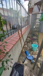 Allée extérieure avec clôture en fer, balais, seau turquoise et échelle verte. Une Fiat 462-NX est garée dans la rue derrière.