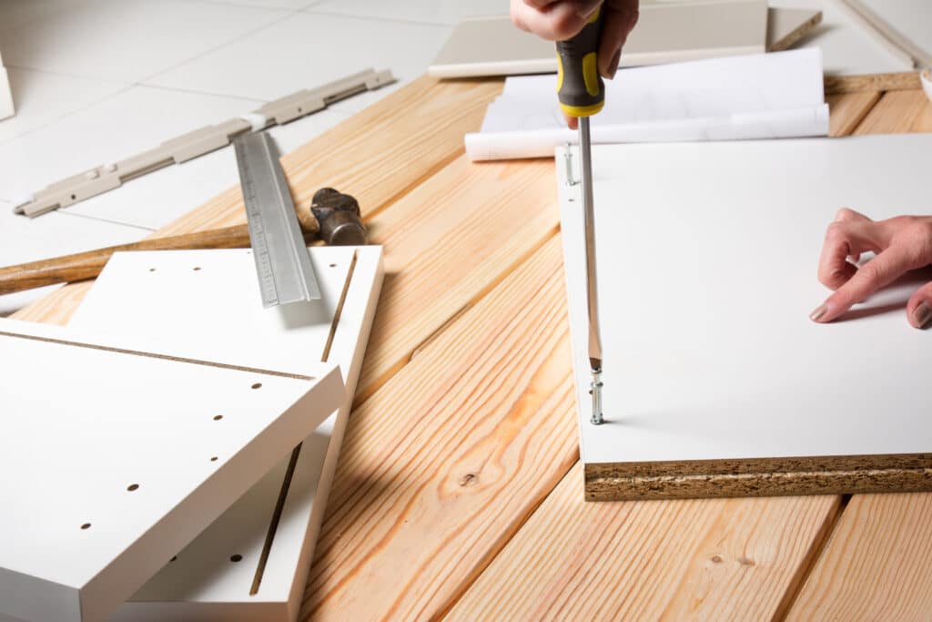 Personne assemble un meuble en kit, vissant une planche blanche en aggloméré avec un tournevis. Outils et plans sur parquet clair.