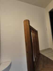 Dossier et assise d'une chaise en bois clair, contre un mur lumineux. On aperçoit un couloir avec carrelage et interrupteurs muraux.