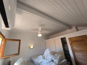 Chambre lumineuse avec plafond lambrissé blanc, climatisation, ventilateur de plafond et lit défait. Applique murale et placards intégrés.