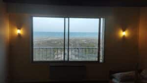 Chambre chaleureuse vue mer: fenêtre sur plage, végétation, vagues calmes. Deux appliques murales allumées, lit visible.