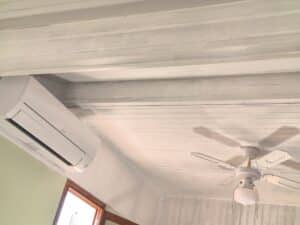 Plafond en bois blanc rainuré avec climatiseur mural Mitsubishi Electric et ventilateur de plafond. Mur vert et cadre de fenêtre en bois.