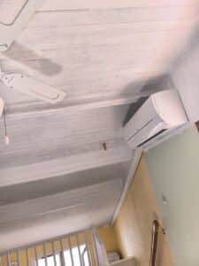 Plafond en lattes de bois blanc, ventilateur de plafond, climatisation murale. Murs jaune et vert avec détails bois.