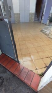 Cour extérieure carrelée beige avec taches d'humidité, vue depuis un seuil rouge. Un portail gris ouvert, des portes et un escalier.