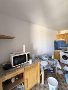 Cuisine en rénovation désordonnée avec micro-ondes blanc, machine à laver, outils, bouteilles et seau de peinture. Comptoirs et carrelage tachés.