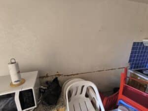 Cuisine avec mur beige endommagé par une fissure horizontale, montrant un micro-ondes, un rouleau essuie-tout et des chaises empilées. Un carrelage mural bleu et des outils de chantier sont visibles.