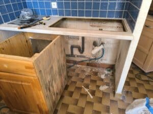 Cuisine en rénovation avec évier retiré, comptoir marbré, plaque de cuisson et outils (NUSSBAUM). Carrelage mural bleu, plomberie apparente, sol carrelé sale.