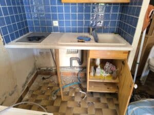 Cuisine en désordre : carrelage bleu, évier, plaques. Murs abîmés, plomberie visible, sol sale. Meuble sous-évier ouvert, produits 'MAXIPACK ANTI-BACTERIEN'.