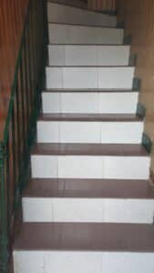 Escalier intérieur carrelé blanc et marron, avec main courante verte, montant le long d'un mur en bois. Contremarches claires, marches foncées anti-dérapantes.