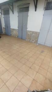 Façade blanche avec soubassement en pierre, trois portes doubles grises et fenêtres à barreaux. Sol carrelé beige extérieur.