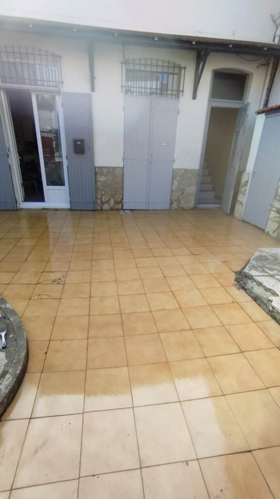 Patio extérieur carrelé beige et humide devant une façade de maison blanche. Porte vitrée ouverte, grande porte grise, et escalier intérieur. Carrelage fissuré.