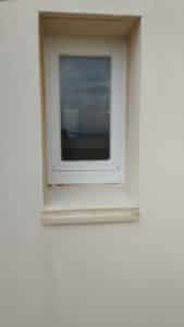 Fenêtre blanche rectangulaire dans un mur texturé beige clair, son verre reflète un ciel gris nuageux, la mer lointaine et des fils électriques.