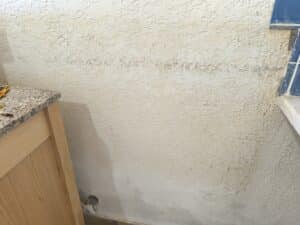 Mur beige avec peinture écaillée et cloquée due à l'humidité. Des taches de dégât des eaux sont visibles, près d'un meuble en bois clair et d'un comptoir en granit.