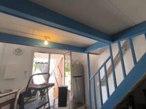 Intérieur lumineux avec poutres et rampe d'escalier bleues. Horloge murale, ampoule allumée, portes vitrées, échelle, frigo Continental Edison.