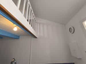 Intérieur blanc : mezzanine avec balustrade bois et dessous bleu. Ampoule lumineuse pend, horloge murale blanche (2h40) et meuble bas.