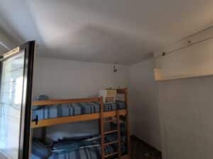 Chambre avec lits superposés bleus et blancs, fenêtre à gauche, crochet mural, meuble de rangement blanc à droite et sol carrelé.