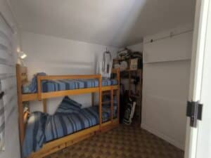 Chambre compacte avec lits superposés en bois et literie bleue rayée. Étagères de rangement encombrées, armoire blanche et sol carrelé.