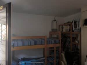 Lits superposés en bois avec literie bleue dans une chambre avec fenêtre lumineuse. Rangement mural IKEA, échelle et chaises empilées.