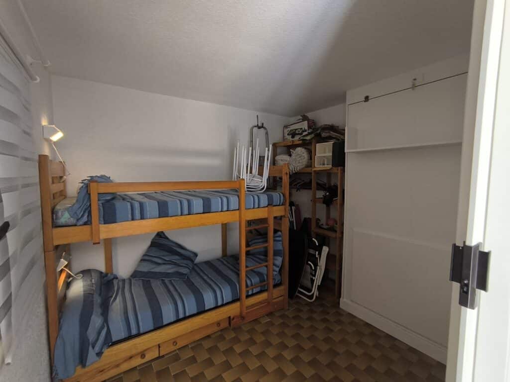Chambre simple avec lits superposés en bois à literie rayée bleue. Étagères de rangement, armoire blanche et sol carrelé marron.