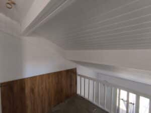 Loft lumineux avec plafond et murs lambrissés blancs. Mur accent bois et balustrade blanche. Vue sur un espace inférieur éclairé.