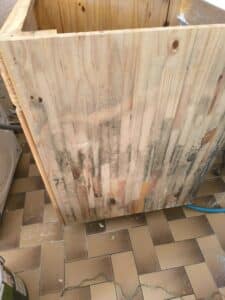 Meuble en bois clair non traité, fortement moisi et taché de rouge, posé sur un sol carrelé. Tuyau bleu, prise électrique et câbles au sol.