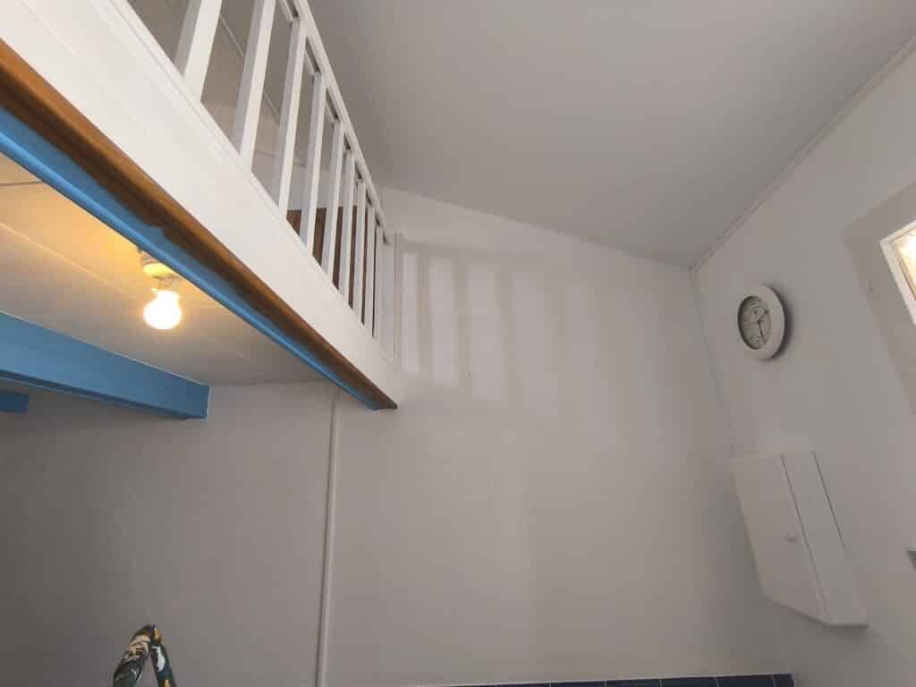 Intérieur lumineux: mezzanine blanche, rampe bois et bleu. Poutres bleues, ampoule. Horloge murale (10h) sur mur blanc. Travaux de rénovation.