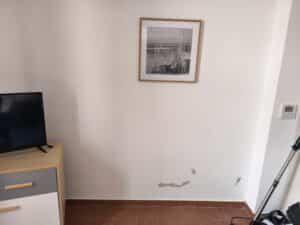 Pièce lumineuse au mur blanc, peinture écaillée visible au bas. TV sur commode bois/gris. Cadre noir et blanc de bateaux. Thermostat Daikin 20°C.