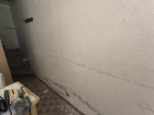 Mur texturé beige avec dégâts d'humidité et fissure linéaire au centre. Ponceuse Dewalt sur table carrelée, escalier sombre en arrière-plan.