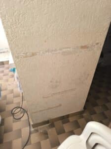 Mur beige texturé avec peinture écaillée et zones dénudées, indiquant des dommages. Sol carrelé marron et un câble noir en bas.