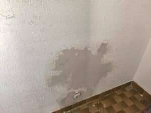 Mur blanc texturé avec grande zone de peinture écaillée, exposant l'enduit gris. Une prise électrique est visible, sol carrelé marron.