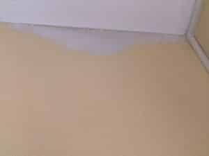 Mur blanc texturé et sol beige avec revêtement texturé. Un joint gris-violet irrégulier est visible à la jonction et un profilé blanc dans le coin.