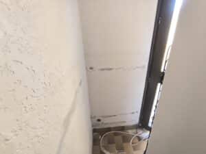 Mur texturé clair avec des taches sombres, un trou et des câbles électriques au sol. Coin d'une pièce avec carrelage et lumière vive d'une porte/fenêtre.