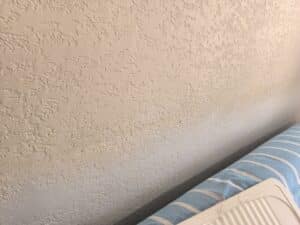 Mur texturé beige clair, en partie ombragé, avec un coussin bleu rayé de blanc et un coin de plateau en plastique crème en bas à droite.