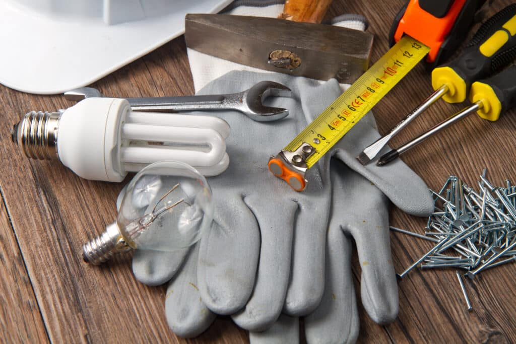 Outils de bricolage sur bois: gants, marteau, mètre ruban (3m, 4m, 5m), tournevis, ampoules (CFL, incandescente), clous. Casque blanc.