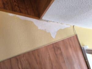 Dégâts de peinture murale jaune texturée écaillée sous un plafond texturé blanc. Boiseries foncées et panneau mural imitation bois.