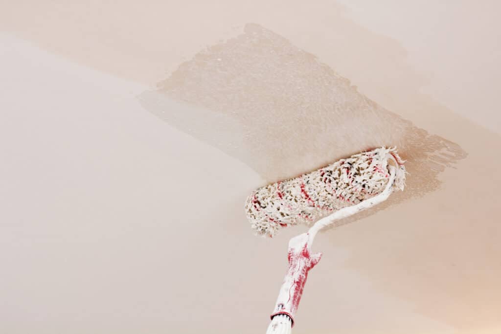 Rouleau de peinture blanc et rouge appliquant de la peinture crème sur un plafond clair. Une section fraîchement peinte est visible.