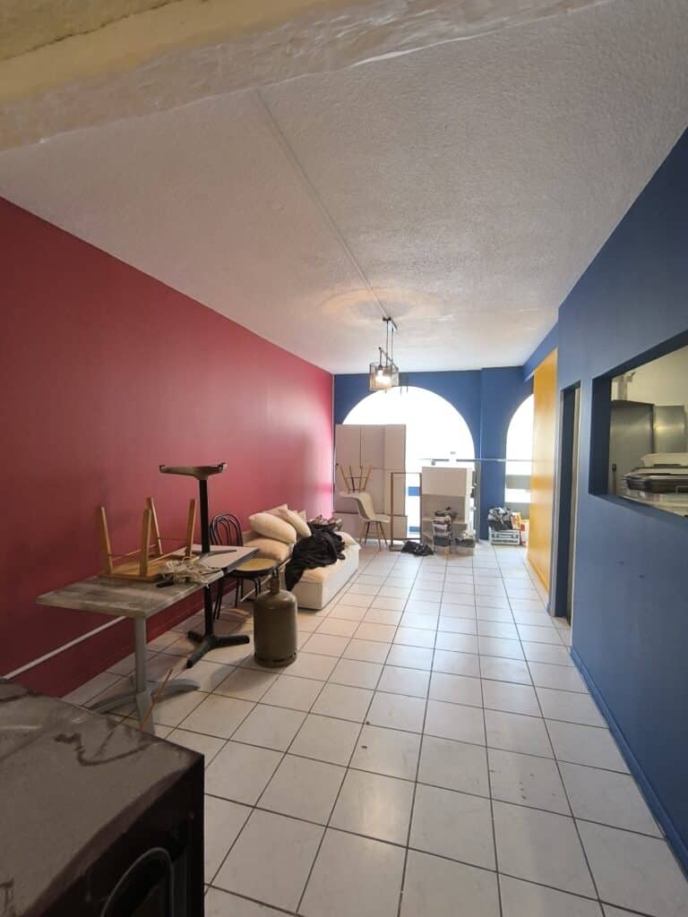 Grande pièce rouge et bleue avec plafond texturé, sol carrelé blanc. Tables, chaises, canapé et bonbonne de gaz. Ouvertures arquées, comptoir bleu.