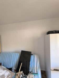 Canapé rayé, TV noire, outils et planche plâtrée dans une pièce en travaux. Un réfrigérateur blanc et un mur texturé occupent l'espace.