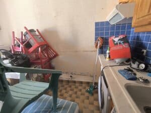 Pièce de service désordonnée avec chaises rouges empilées, mur crème écaillé, carrelage abîmé. Évier, machine à laver, produits Voltair et Ripolin sur le comptoir.