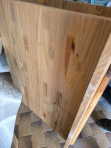 Planches de bois de pin brut avec grain et nœuds, certaines avec des résidus de colle blanche sur les bords, posées sur un sol carrelé marron.