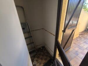 Porte en bois ouverte sur un patio carrelé ensoleillé. Intérieur sombre avec mur écaillé, échelle métallique et appareil blanc, sol à carreaux marron, câbles.