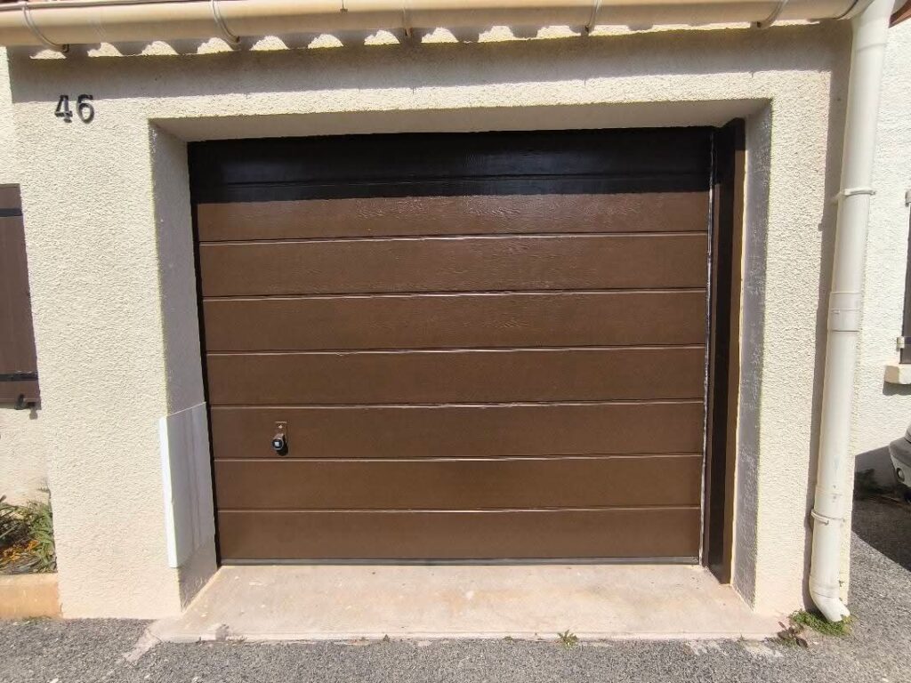 Porte de garage sectionnelle marron horizontale avec numéro 46 sur façade crème. Gouttière et volet visible, allée en gravier.