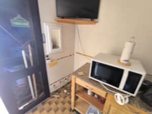 Coin cuisine en rénovation avec micro-ondes sur comptoir, TV sur étagère, porte sombre affichant un logo Heineken, et mur endommagé.