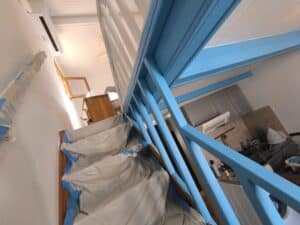Escalier intérieur en bois en cours de peinture, avec une rampe fraîchement peinte en bleu vif. Les marches sont protégées par des bâches. Climatisation et cuisine visible en arrière-plan.