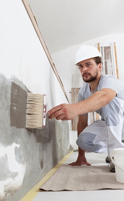 Artisan avec casque blanc appliquant un enduit d'étanchéité gris sur un mur avec un gros pinceau. Chantier de rénovation.