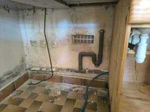 Coin utilitaire ancien et sale : murs à peinture écaillée, traces d'humidité et moisissures. Tuyauterie PVC, prises électriques, grille ventilation et carrelage poussiéreux.