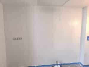 Mur blanc en cours de peinture. Prises électriques, ruban de masquage bleu, rouleau et bac de peinture au sol. Grille de ventilation au plafond.