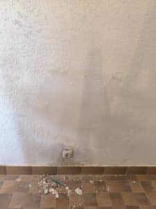 Mur texturé beige en cours de rénovation, avec des zones d'enduit arraché. Une prise électrique est visible au-dessus d'un sol carrelé sale et de débris de chantier.