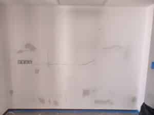 Mur blanc avec fissures, zones de réparations en plâtre gris et prises électriques. Sol protégé par du plastique bleu. Préparation rénovation.